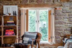 Ein gemütliches Zimmer mit Steinwand, einem hölzernen Bücherregal, einem Fenster mit geöffneten Klappläden und einem Stuhl mit gestreiftem Kissen.
