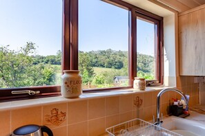 Uma cozinha com vista para uma paisagem verde através de grandes janelas, uma chaleira, uma pia e dois potes rotulados 'FLOUR' e 'SUGAR'.