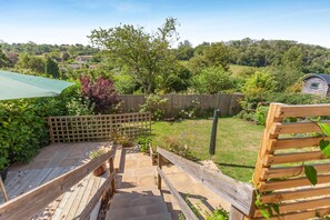 Une terrasse en bois avec un banc en bois, une clôture en bois et une vue sur une cour verdoyante, avec des arbres et une maison en arrière-plan.