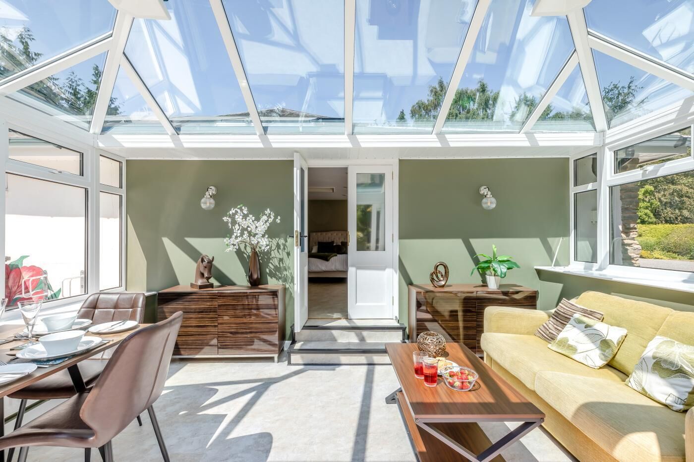 Een modern interieur met een glazen dak, een eethoek, een bank en een keuken die door een open deur zichtbaar is.