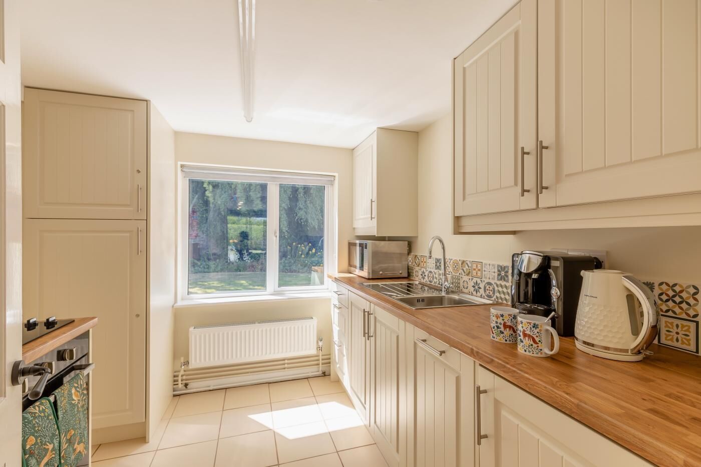 A kitchen with white cabinets, a window, a radiator, a microwave, a kettle, and a coffee maker.