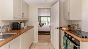 A compact kitchen with white cabinets, a coffee machine, and a sink. A door leads to a bedroom with a bed and a view of greenery. - 1 Bed in Combe (oc-g45502) (Combe)