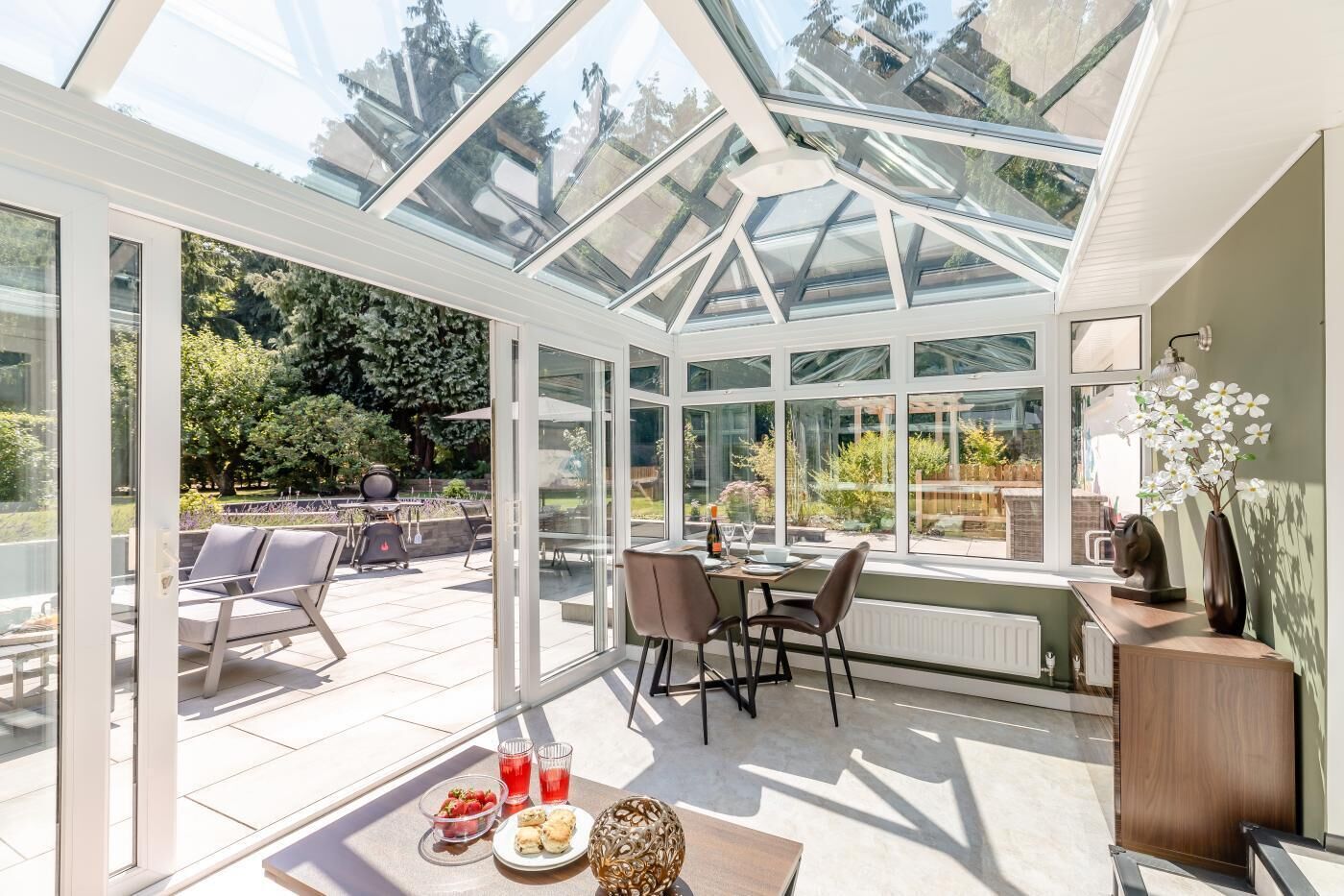 A conservatory with a glass roof, a dining table with chairs, and a view of the garden outside.