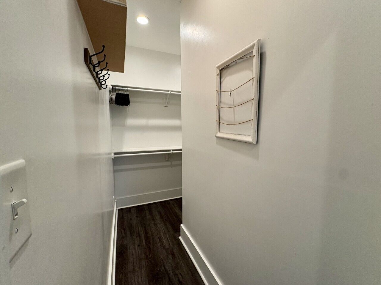 A narrow hallway with white walls, a wooden floor, a coat rack, a window with blinds, and a light switch.