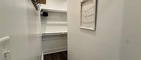 A narrow hallway with white walls, a wooden floor, a coat rack, a window with blinds, and a light switch.