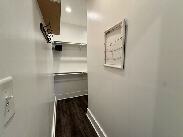 A narrow hallway with white walls, a wooden floor, a coat rack, a window with blinds, and a light switch.