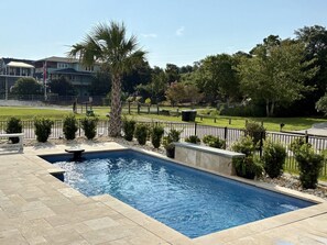 A rectangular swimming pool with a stone border, surrounded by a low fence, and a palm tree in the background. - Sound view with Private Saltwater Pool - A Tad Salty (Emerald Isle)