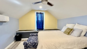 Quarto com cama, um banquinho, ventilador de teto e janela com cortinas.