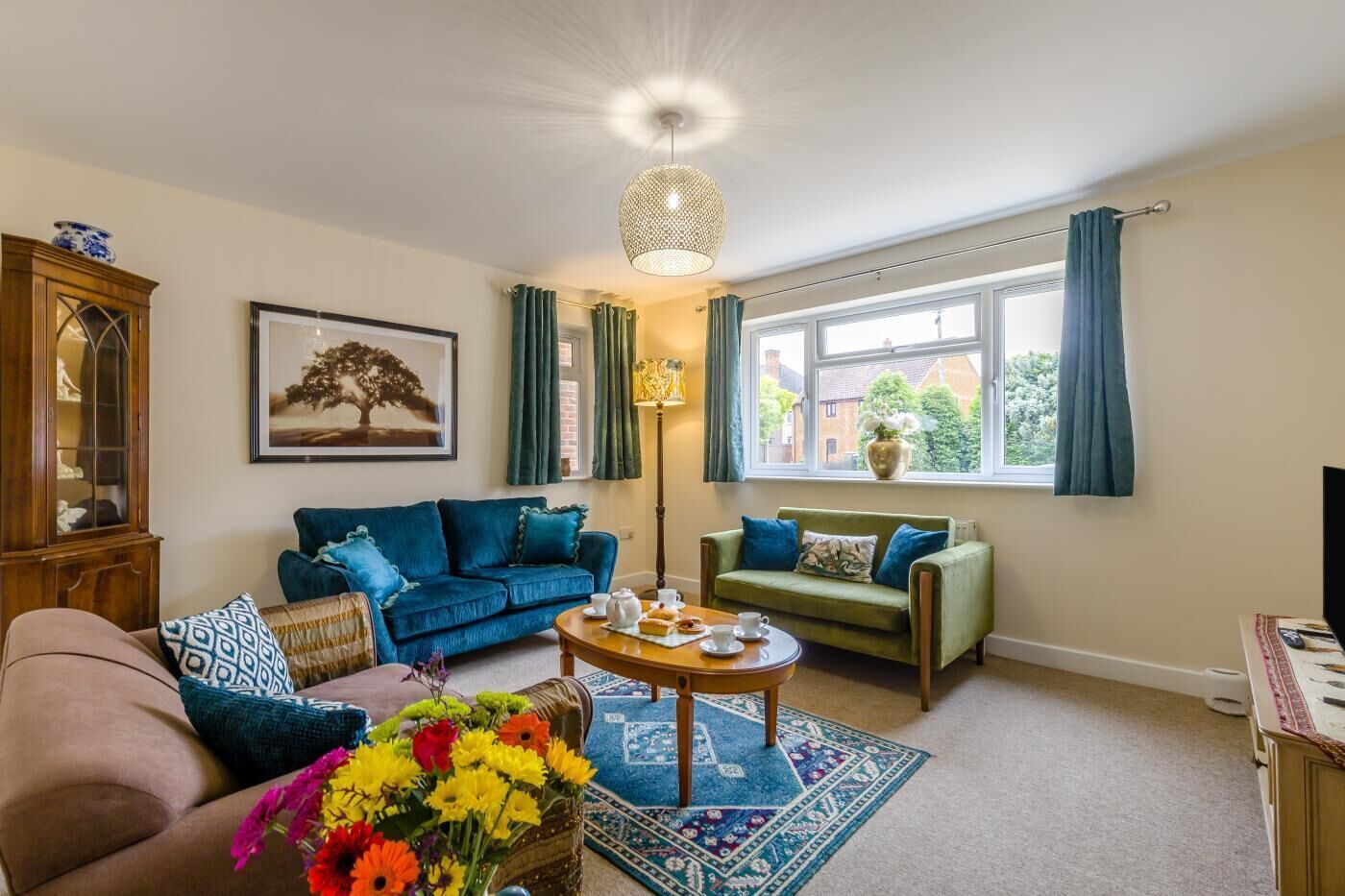 Ein Wohnzimmer mit blauem Sofa, grünem Sofa, Holzschrank, Gemälde, Fenster mit Vorhängen und einer Vase mit Blumen.