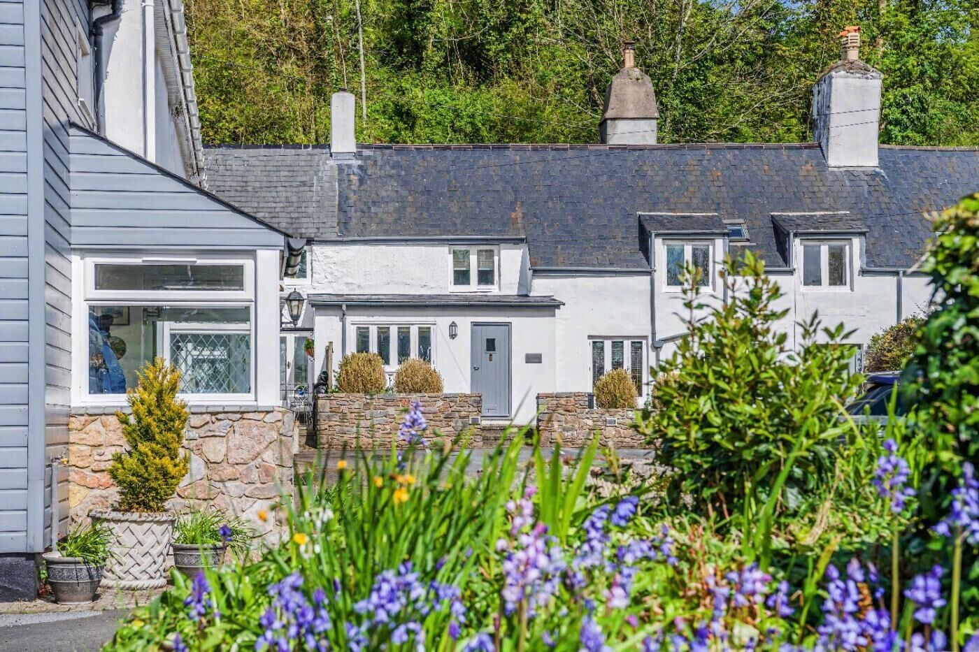 A row of white houses with stone walls and multiple chimneys, surrounded by greenery and flowering plants.
