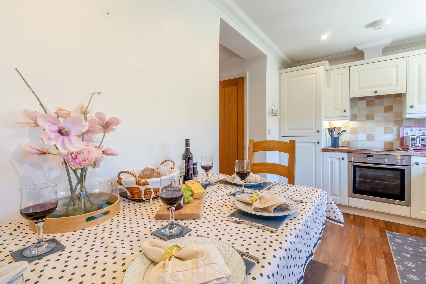 A dining area with a table set for two, featuring a polka dot tablecloth, a vase of flowers, a bottle of wine, and a basket of bread.