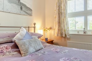 Ein Schlafzimmer mit einem Bett, Kissen, einem Nachttisch, einer Vase mit Blumen und einem Fenster mit Vorhängen.