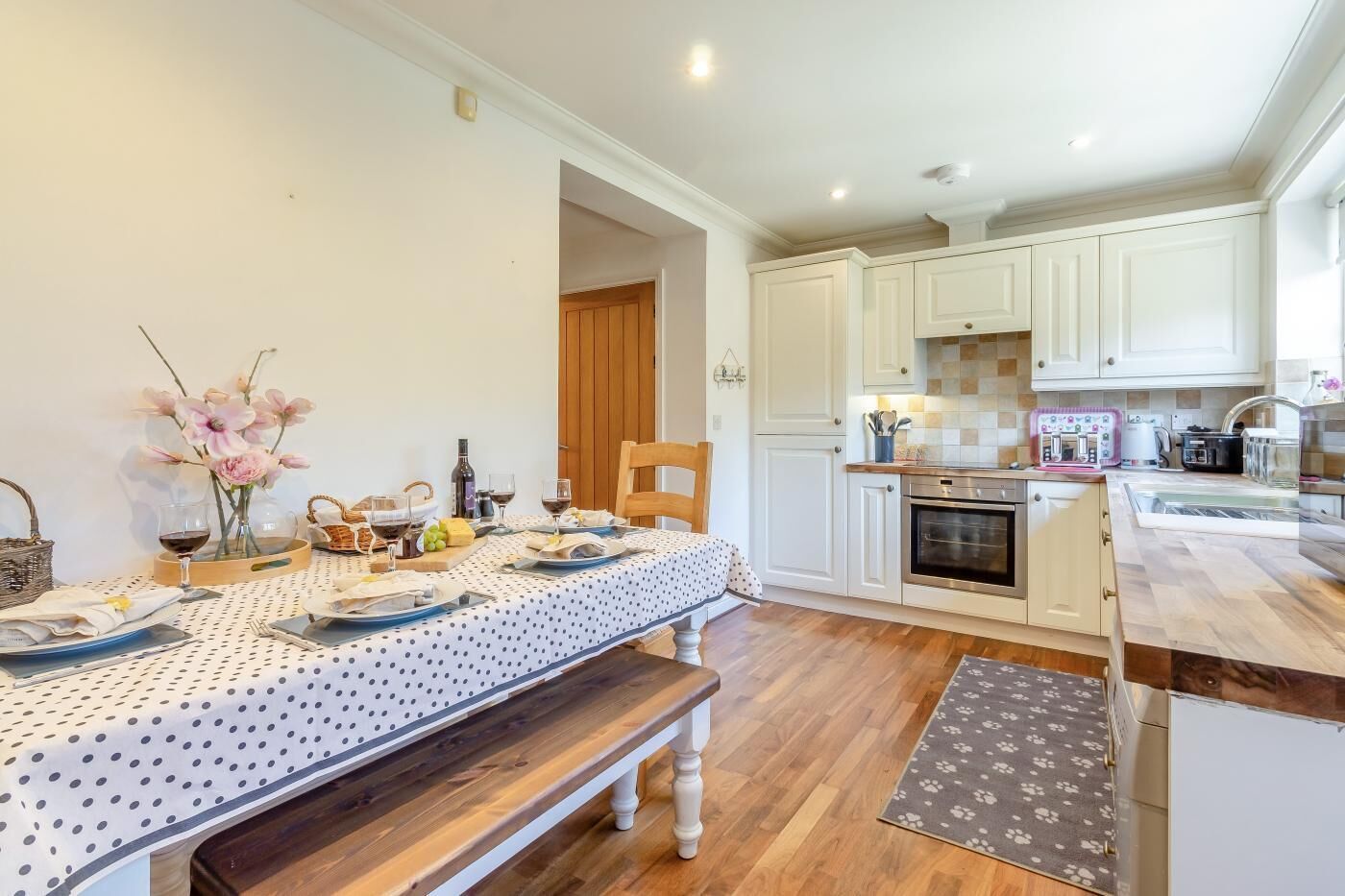 A dining area with a table set for two, a bench, and a kitchen with white cabinets and a wooden countertop.