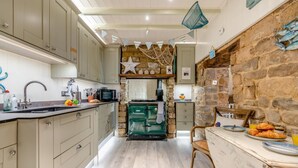 A kitchen with a stone wall, a stove, and a dining area with a table and chairs. - 3 Bed in Robin Hoods Bay, Whitby (oc-g43284) (Robin Hood's Bay)