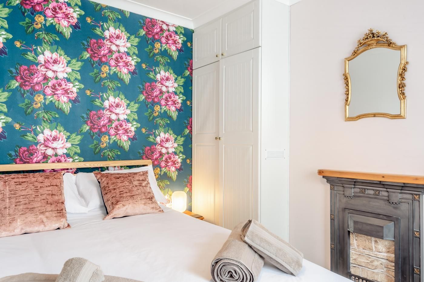 A hotel room with a bed, a mirror, a fireplace, and a floral wallpaper.