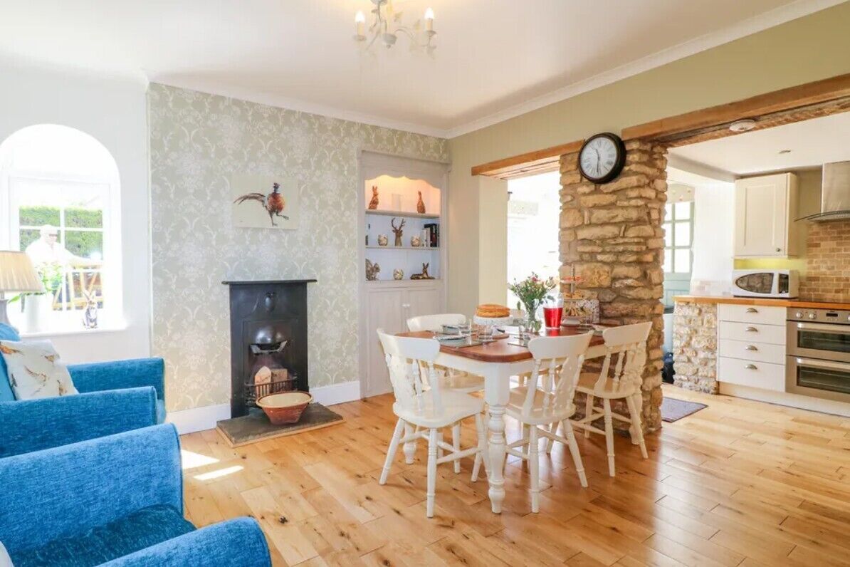A cozy living room with a fireplace, a dining area with white chairs, and a kitchen with a stone wall and modern appliances.