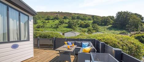 Une terrasse dotée de mobilier d’extérieur, offrant une vue sur un paysage vallonné, et un bâtiment avec de grandes fenêtres.
