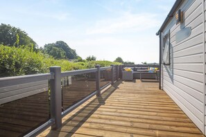 A wooden deck with a railing, a sofa, and a table, overlooking a garden with trees and a building. - 3 Bed in Abersoch (oc-a45542) (Abersoch)