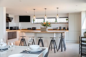 A modern kitchen with a central island, bar stools, and a dining area with a table set for two. - 7 Bed in Broadwoodkelly (oc-b45447) (Broadwood Kelly)
