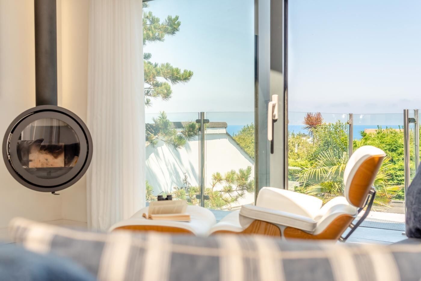 A room with a large window, a circular mirror, a chair with a book, and a view of the sea.