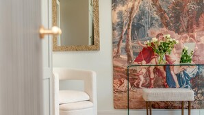 Et værelse med en hvid lænestol, et sofabord med glasplade og en vase med blomster, samt et stort maleri i klassisk stil på væggen.