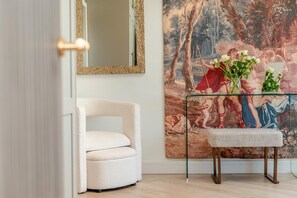 Ein Zimmer mit einem weißen Sessel, einem Glastisch mit einer Blumenvase und einem großen Gemälde im klassischen Stil an der Wand.