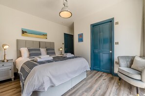 Ein Hotelzimmer mit Bett, Nachttischlampen, einer blauen Tür, einem grauen Sofa und Bildern an den Wänden.