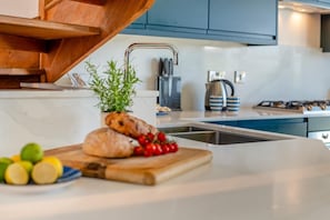 Eine Küche mit einem Holzkörperschen, einem Spülbecken und einer Arbeitsplatte mit Schneidebrett, auf dem Brot und Tomaten liegen.