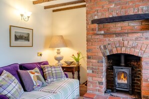 A cozy living room with a brick fireplace, a sofa with plaid cushions, a table with a lamp, and a framed picture on the wall. - 2 Bed in Castle Acre (oc-r18323) (Castle Acre)