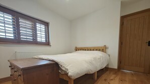 Ein Schlafzimmer mit einem hölzernen Bett, einer Holzkommode, einem Heizkörper, einem Fenster mit Jalousien und einer Holztür.