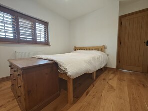 Ein Schlafzimmer mit einem hölzernen Bett, einer Holzkommode, einem Heizkörper, einem Fenster mit Jalousien und einer Holztür.