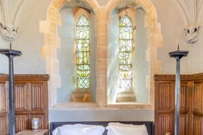 Una habitación con dos amplios ventanales de vitral, paneles de madera y una cama con ropa blanca.
