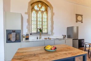 Una cocina con una mesa de madera, un bol de fruta, un ventanal con vidrieras y electrodomésticos modernos.