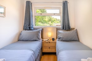 A room with two single beds, a nightstand with a lamp and a book, and a window with curtains.