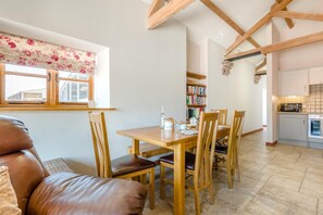 A dining area with a wooden table and chairs, a window with floral curtains, and a kitchen in the background. - 2 Bed in Awre (oc-o45277) (Awre)