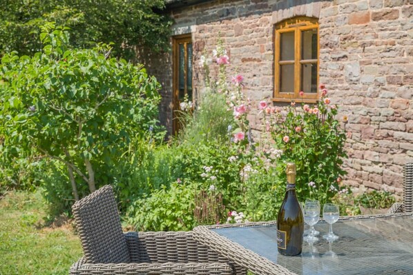 A wicker table with a bottle of wine and glasses, set against a stone building with a window and blooming flowers.