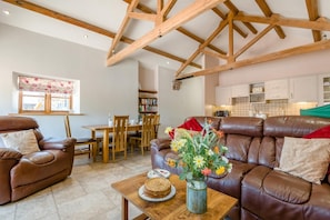 A living room with a brown leather sofa, a dining table with chairs, a bookshelf, and a kitchen area in the background.