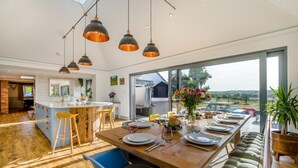 A modern kitchen with a central island, bar stools, and a dining area with a long table set for a meal. - 5 Bed in Marhamchurch (oc-p45735) (Marhamchurch)