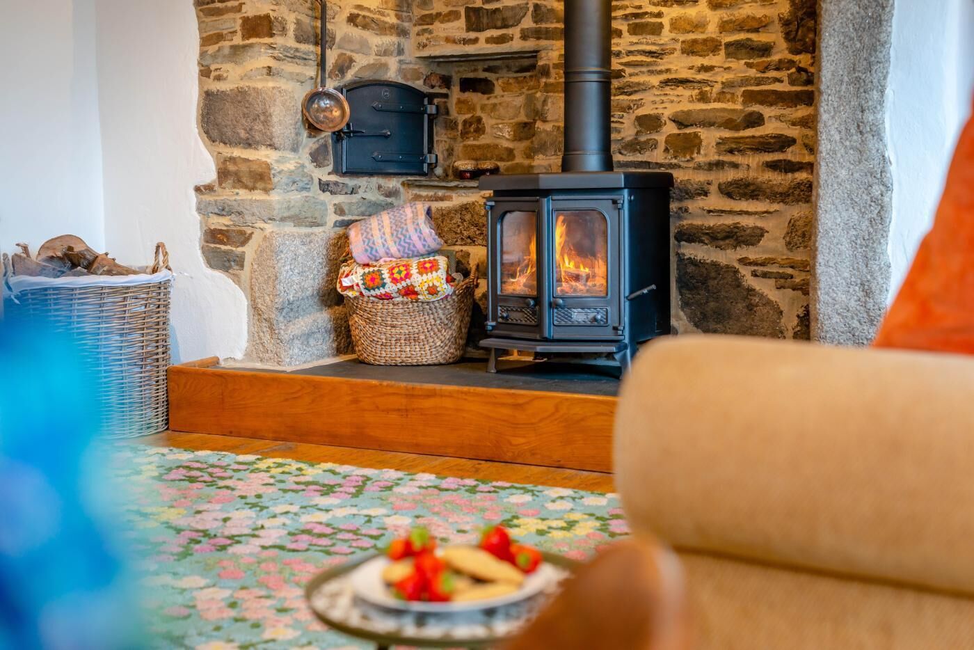 Una habitación acogedora con pared de piedra, estufa de leña, cesta de mimbre y alfombra estampada con motivos florales.