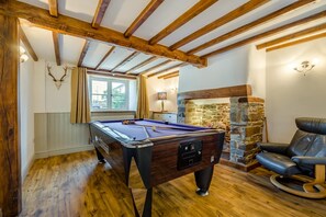 Ein Billardtisch in einem Raum mit Holzbalken, einem Kamin aus Stein und einem Ledersessel.