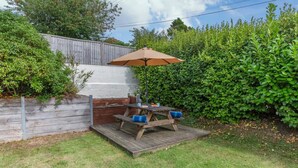 En træterrasse med et picnicbord og parasol, omgivet af et træhegn og frodig vegetation.
