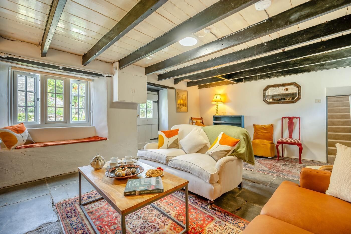 A cozy living room with a sofa, armchairs, a coffee table, and a rug. There is a window with a view of greenery outside.