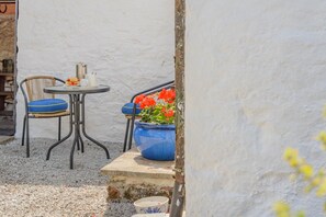 Un petit espace extérieur aménagé avec une table ronde et deux chaises, un pot de fleurs bleu et un mur blanc en arrière-plan.