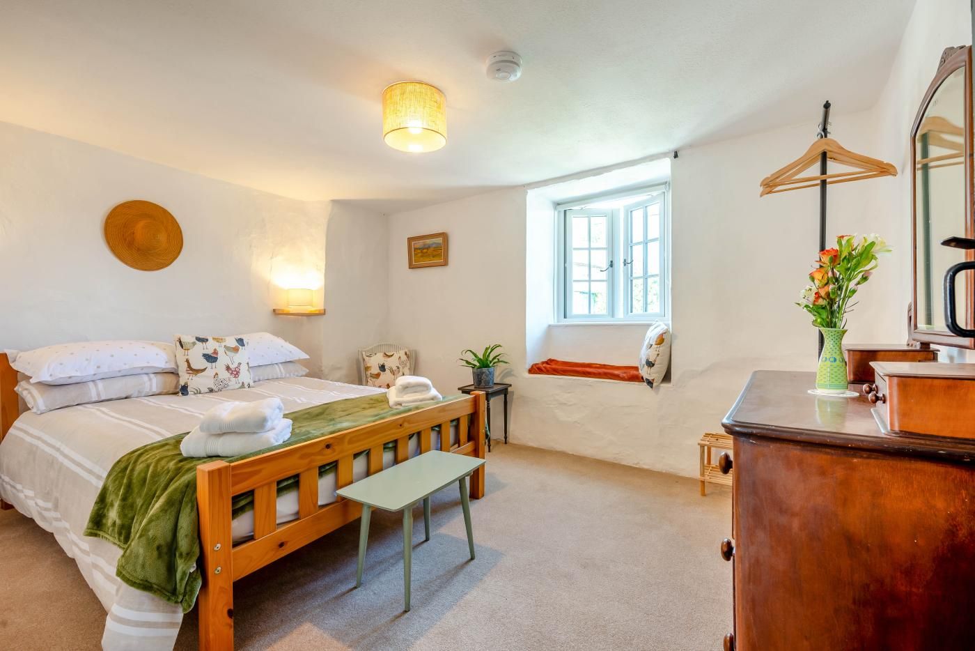 A bedroom with a bed, bedside table, chair, dresser, mirror, window, and a hanging coat rack.