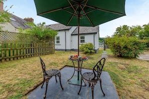 A patio with two chairs and a table under a green umbrella, a wooden fence, and a white building in the background. - 1 Bed in St. Dogmaels (oc-m45322) (Saint Dogmaels)