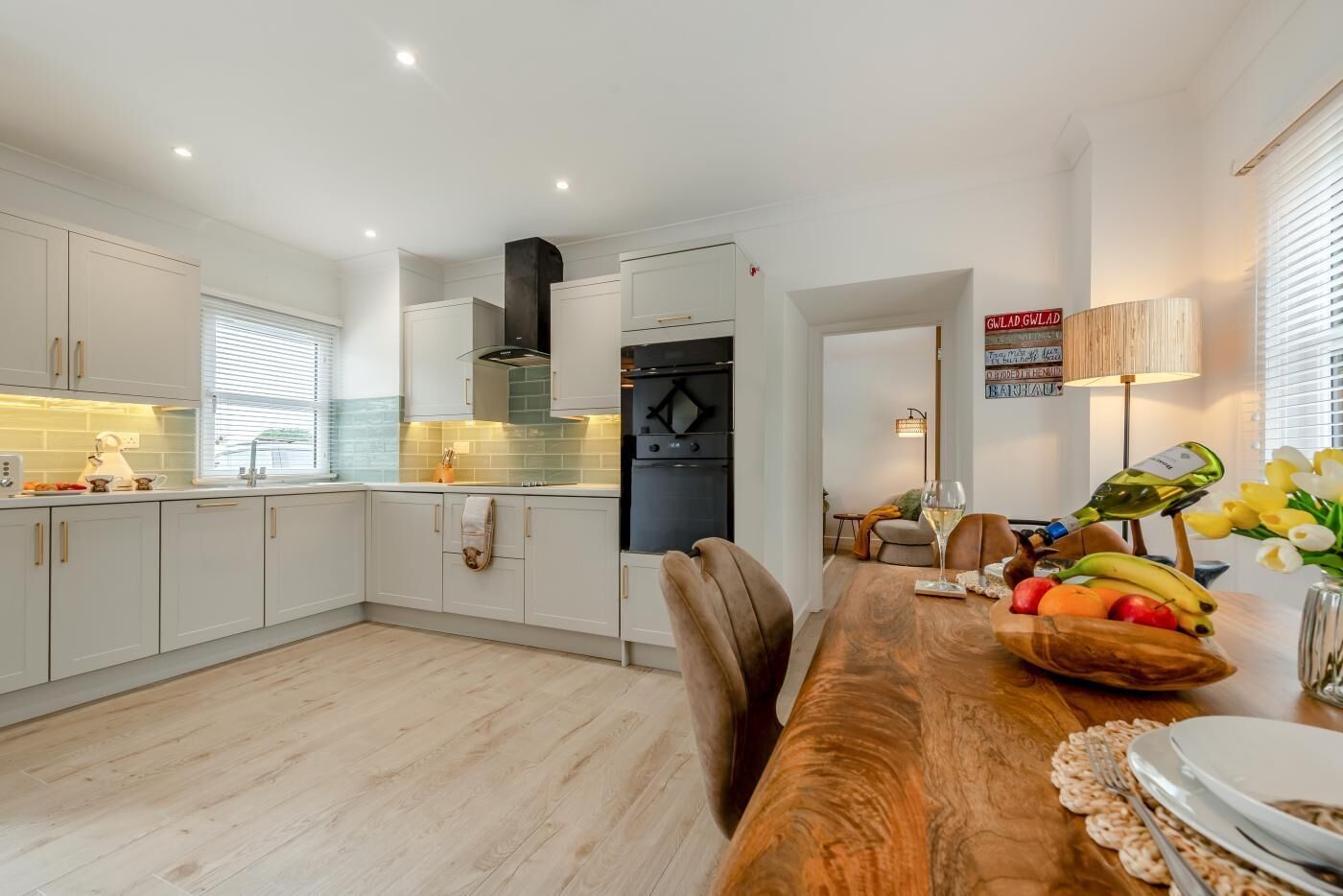 A modern kitchen with white cabinets, a wooden dining table set for two, and a fruit bowl on the table.