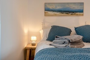 A bed with a blue blanket, pillows, and a folded towel. There is a painting of a beach scene on the wall above the bed. - 1 Bed in St. Dogmaels (oc-m45322) (Saint Dogmaels)