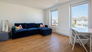 A modern living room with a dark blue sectional sofa, a white dining table, and large windows offering a view of residential buildings. - 3 Bed in Rustington (oc-p45430) (Rustington)