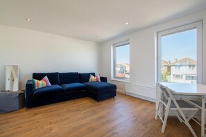 Ein modernes Wohnzimmer mit einem dunkelblauen Ecksofa, einem weißen Esstisch und großen Fenstern mit Blick auf Wohnhäuser.