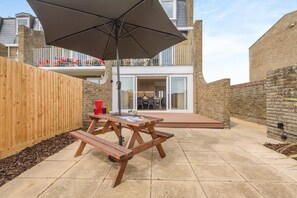 An outdoor patio with a wooden table and benches, a large umbrella, and a view of a residential building with a balcony. - 3 Bed in Rustington (oc-p45430) (Rustington)